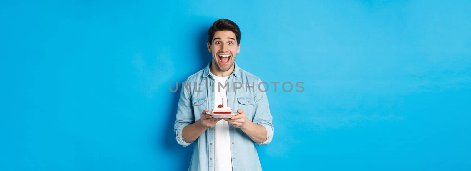 Excited guy holding birthday cake and smiling at camera, celebrating b-day over blue background.