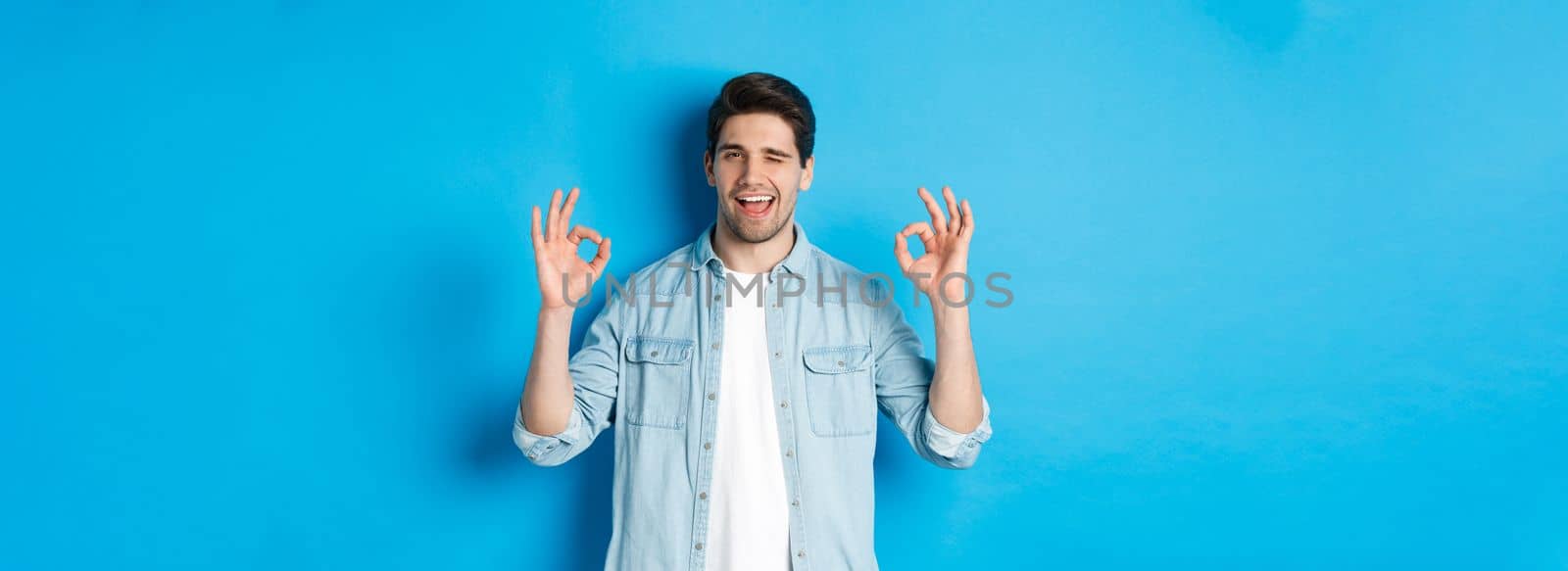 Relaxed and confident man showing ok signs and winking, everything okay gesture, standing against blue background.