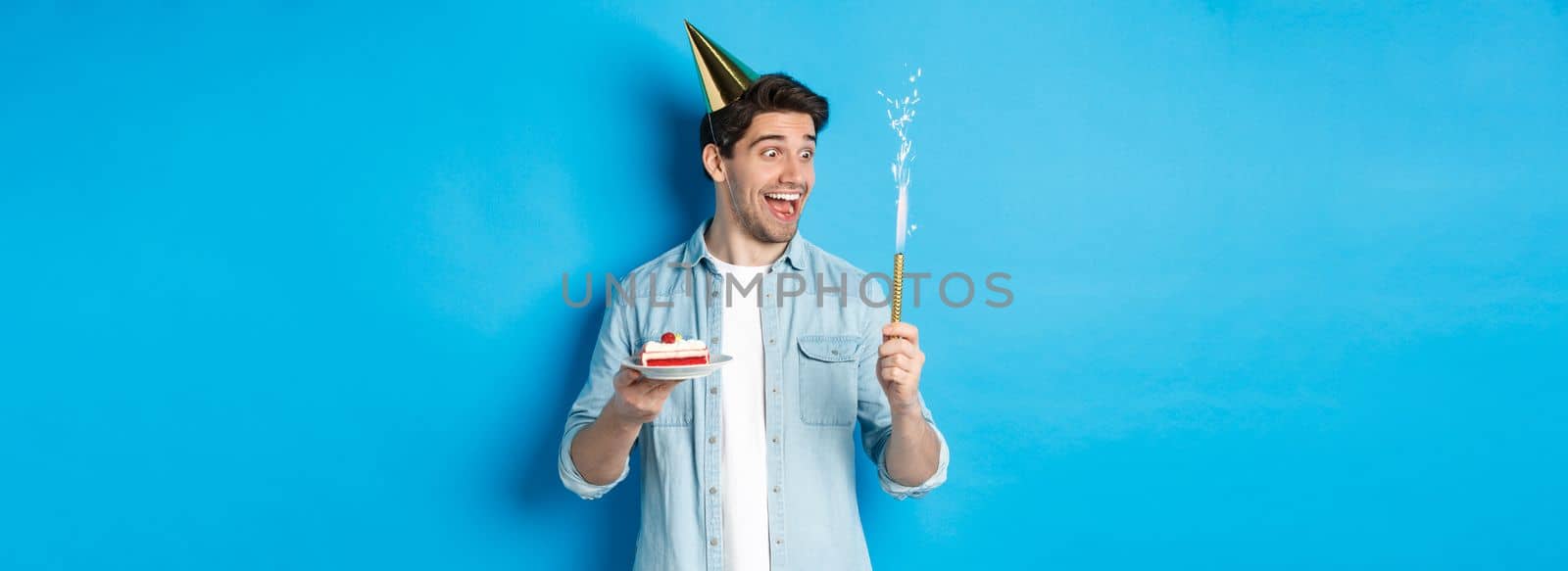 Happy young man celebrating birthday in party hat, holding b-day cake and smiling, standing over blue background by Benzoix