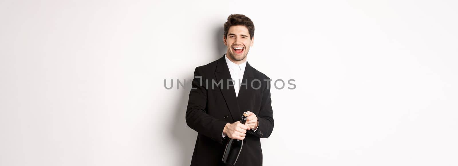 Image of handsome, confident man in black suit, celebrating holiday, open a bottle of champagne and partying, standing joyful against white background by Benzoix