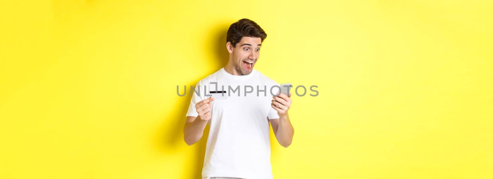 Man looking surprised at smartphone, shopping online, holding credit card, standing over yellow background by Benzoix