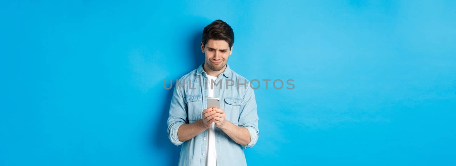 Disappointed and skeptical guy looking at phone, standing over blue background.