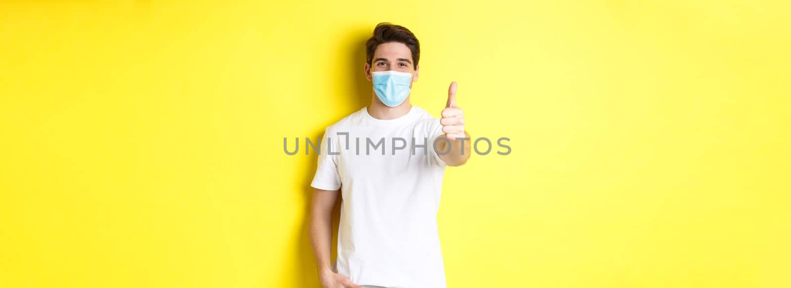 Concept of coronavirus, pandemic and social distancing. Confident young man in medical mask showing thumbs up, yellow background.