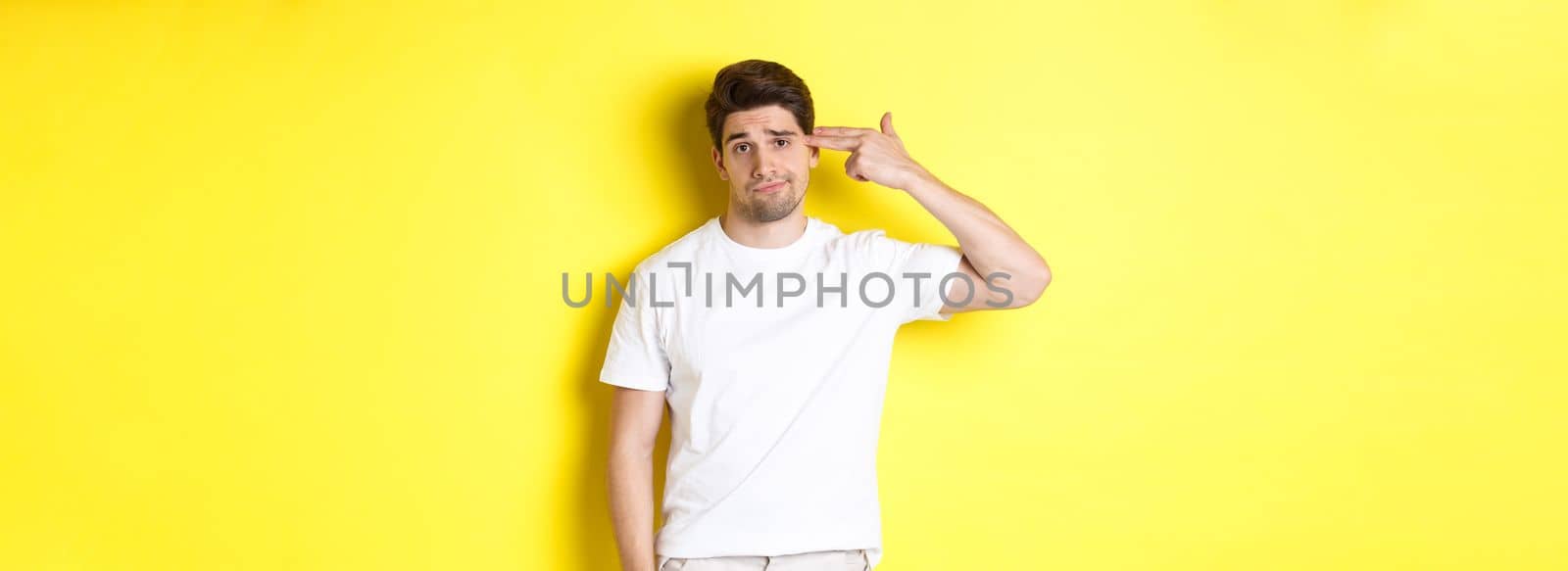 Bothered man shooting himself with finger gun, looking displeased and tired, standing against yellow background.