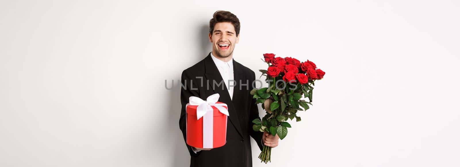 Concept of holidays, relationship and celebration. Charming young man in black suit, holding gift box and bouquet of roses, winking and smiling, standing against white background.