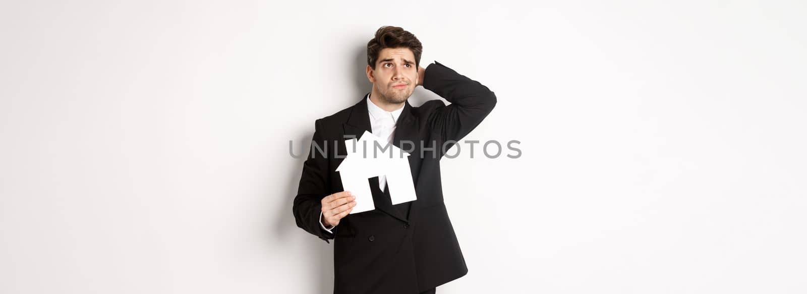 Image of indecisive businessman thinking, holding house maket and looking at upper left corner doubtful, standing against white background.