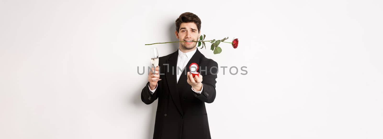 Romantic young man in suit making a proposal, holding rose in teeth and glass of champagne, showing engagement ring, asking to marry him, standing against white background by Benzoix