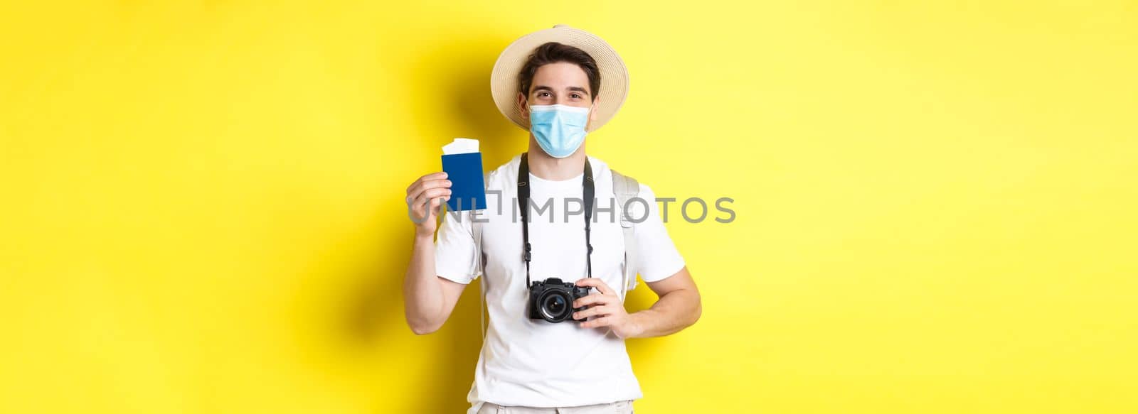 Concept of covid-19, travelling and quarantine. Happy man tourist with camera, showing passport and tickets for vacation, going on trip during pandemic, yellow background.