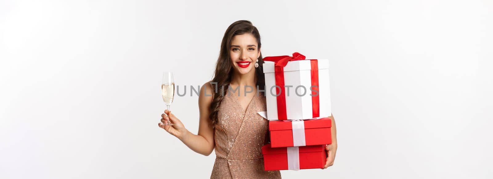 New Year, Christmas and celebration concept. Elegant woman in luxury dress and red lipstick, holding gifts and drinking champagne on party, standing over white background.