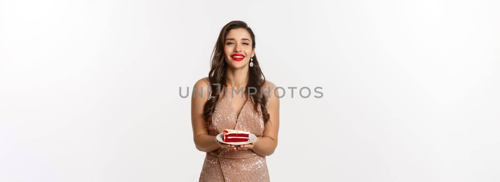 Party and celebration concept. Attractive woman in elegant dress holding delicious cake and smiling with temptation, standing over white background.