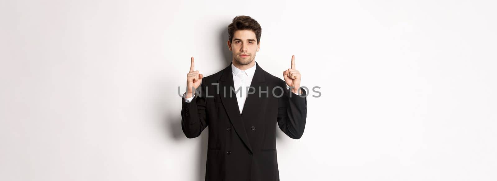 Image of confident and handsome man in formal suit, pointing fingers up, showing copy space on white background.