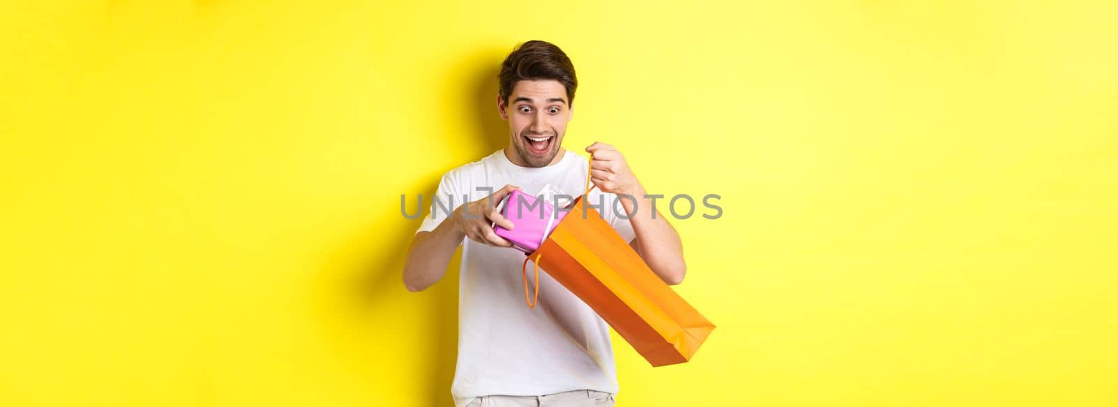 Concept of holidays and celebration. Young man looking surprised as take out gift from shopping bag, standing over yellow background by Benzoix