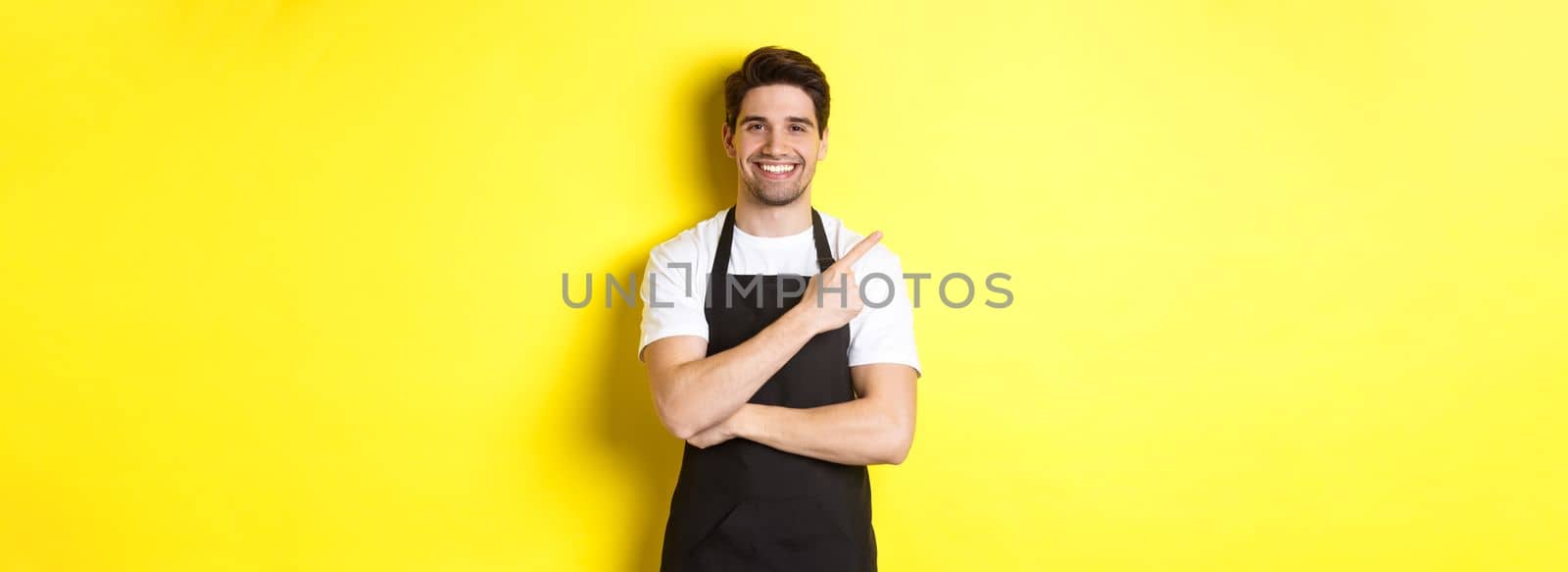 Happy barista pointing finger left and smiling, wearing black apron uniform, standing against yellow background. Copy space