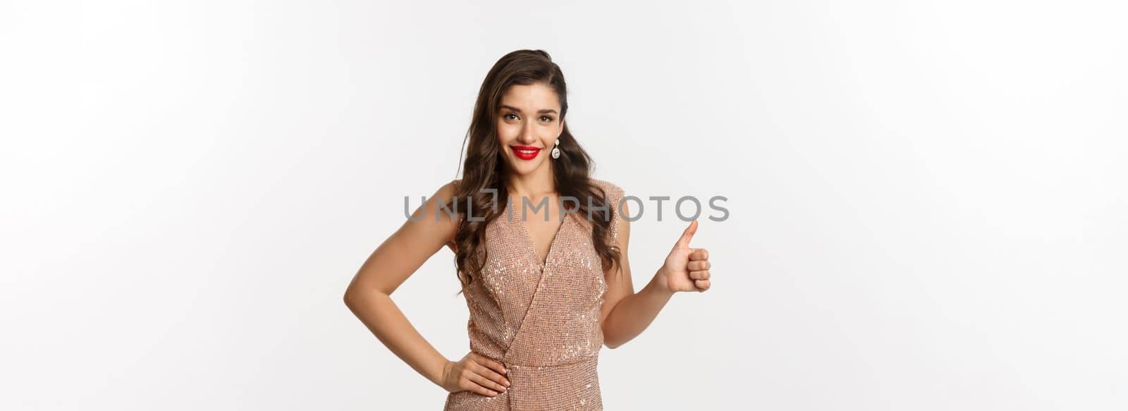 Concept of celebration, holidays and party. Attractive and confident woman with red lips, wearing luxury evening dress and showing thumb up, approve and like, white background.