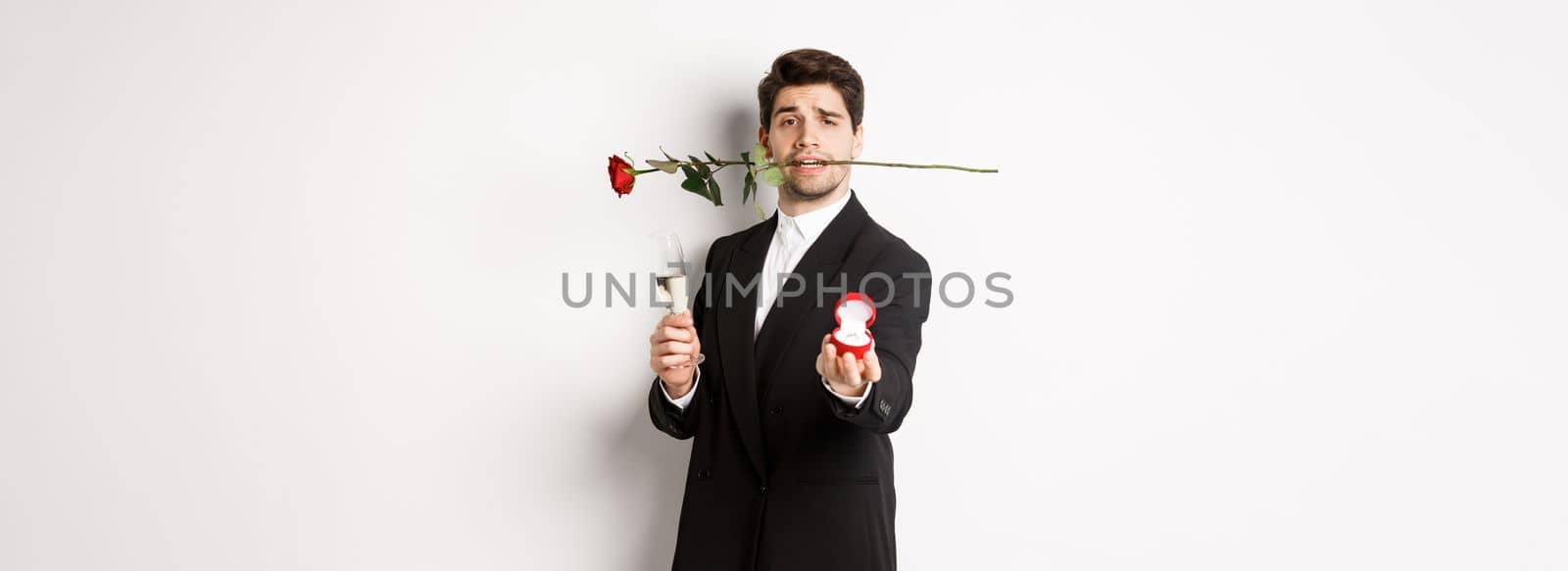 Romantic young man in suit making a proposal, holding rose in teeth and glass of champagne, showing engagement ring, asking to marry him, standing against white background by Benzoix