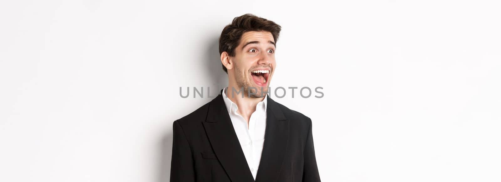 Close-up of handsome male entrepreneur in suit, looking left and smiling amazed, standing against white background.