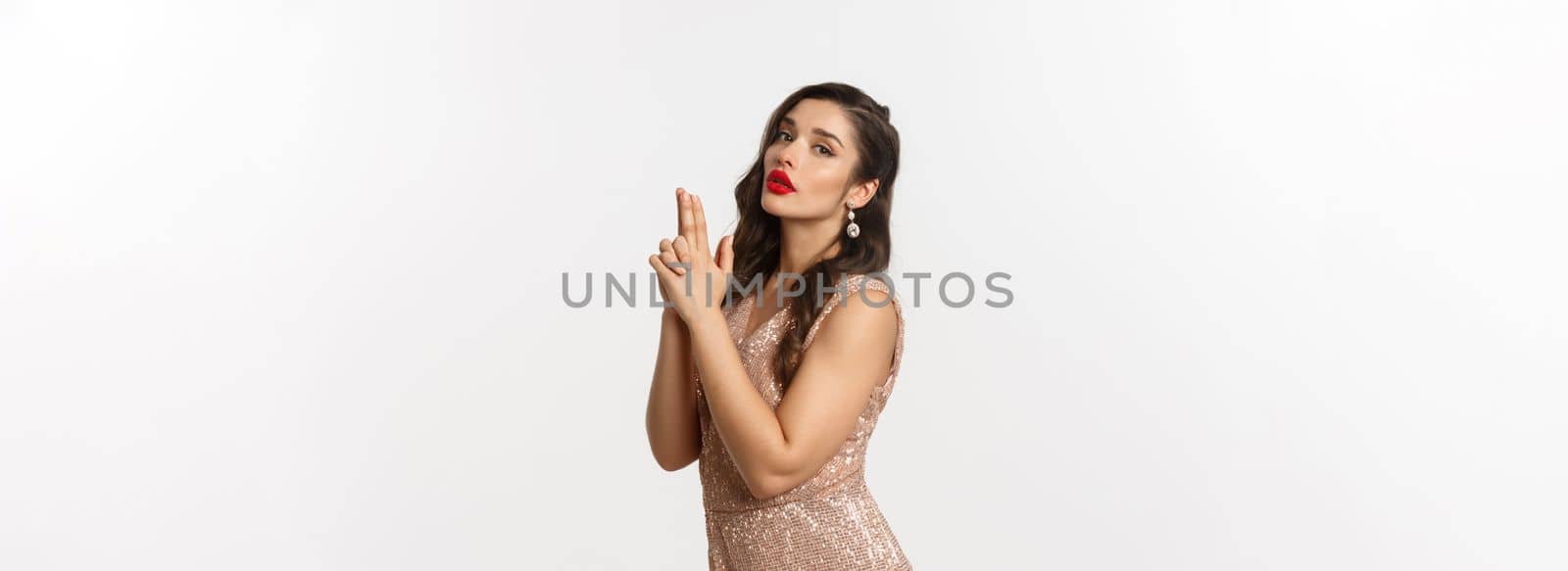 Concept of casino, celebration and party. Sexy woman in glamour dress, red lips, making finger gun pistol gesture and looking at camera, standing over white background.