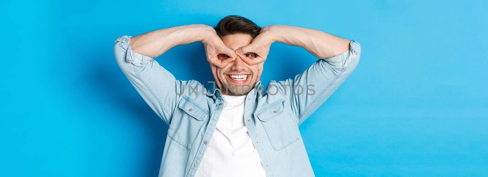 Young caucasian guy showing funny expression, making superhero mask with fingers on eyes, smiling happy, standing over blue background.