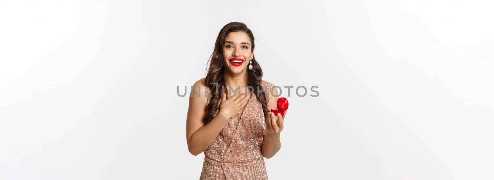 Image of beautiful woman in glamour dress receiving engagement ring, looking surprised and happy at camera, being proposed, standing over white background.