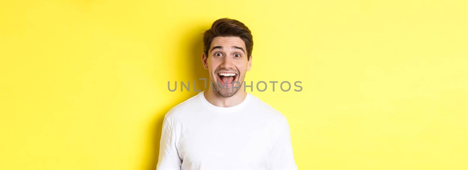 Close-up of surprised handsome guy reacting to great news, standing over yellow background in white t-shirt.