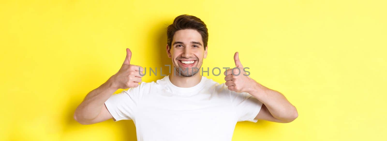Close-up of handsome young man showing thumbs up, approve and agree, smiling satisfied, standing over yellow background by Benzoix