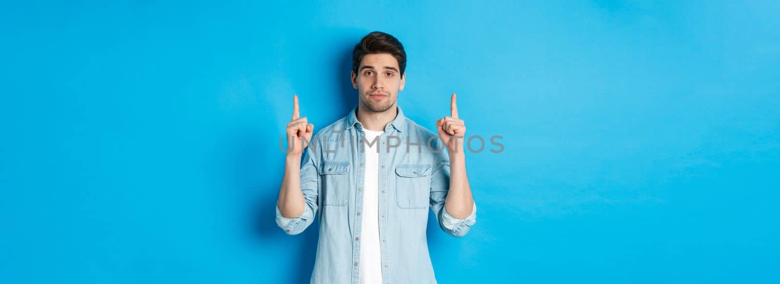 Image of calm handsome man showing you promo offer, pointing fingers up at copy space, standing against blue background.