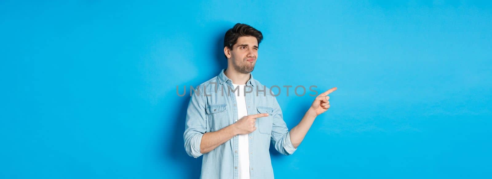 Doubtful adult man pointing fingers left at promotion and looking unsure, grimacing disappointed, standing against blue background.