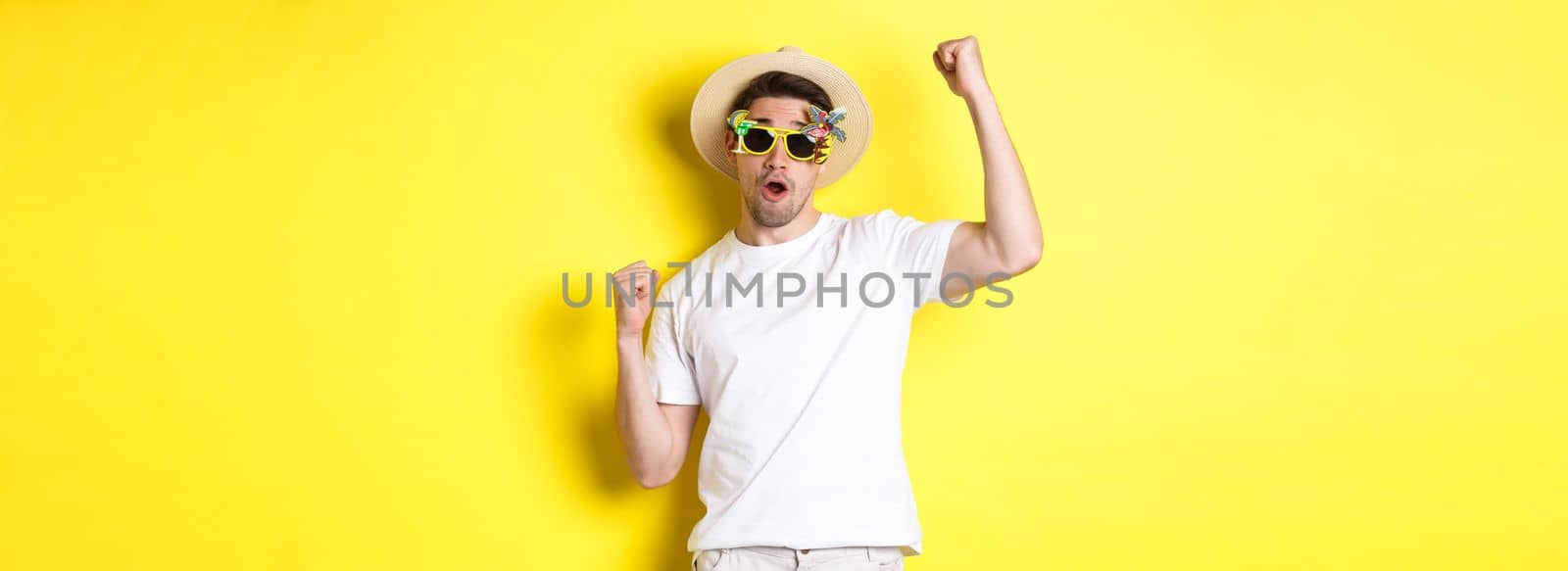 Concept of tourism and lifestyle. Happy guy tourist enjoying trip, rooting for you, fist pump and triumphing, going on journey in summer hat and sunglasses, yellow background.