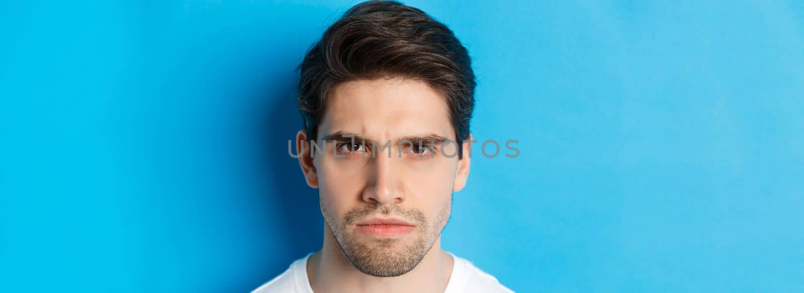 Headshot of angry man frowning, looking disappointed and bothered, standing over blue background.