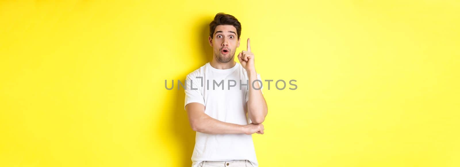 Thoughtful guy suggesting solution, raising finger in eureka sign and looking excited, have an idea, standing against yellow background.