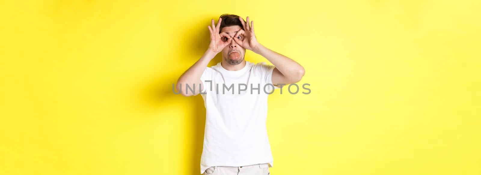 Young man making funny faces and showing tongue, fool around, standing in white t-shirt against yellow background. Copy space