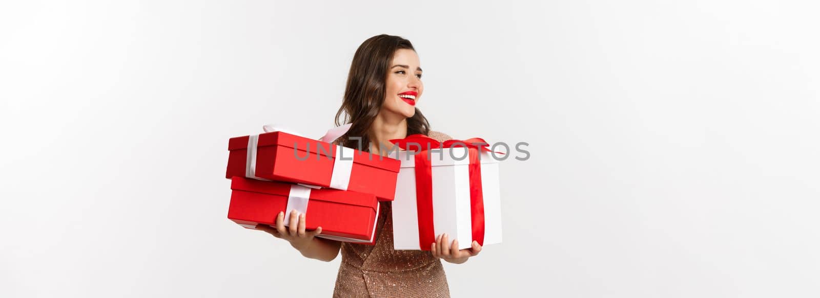 Holidays, celebration concept. Beautiful caucasian woman in elegant dress holding Christmas presents and smiling happy, standing over white background.