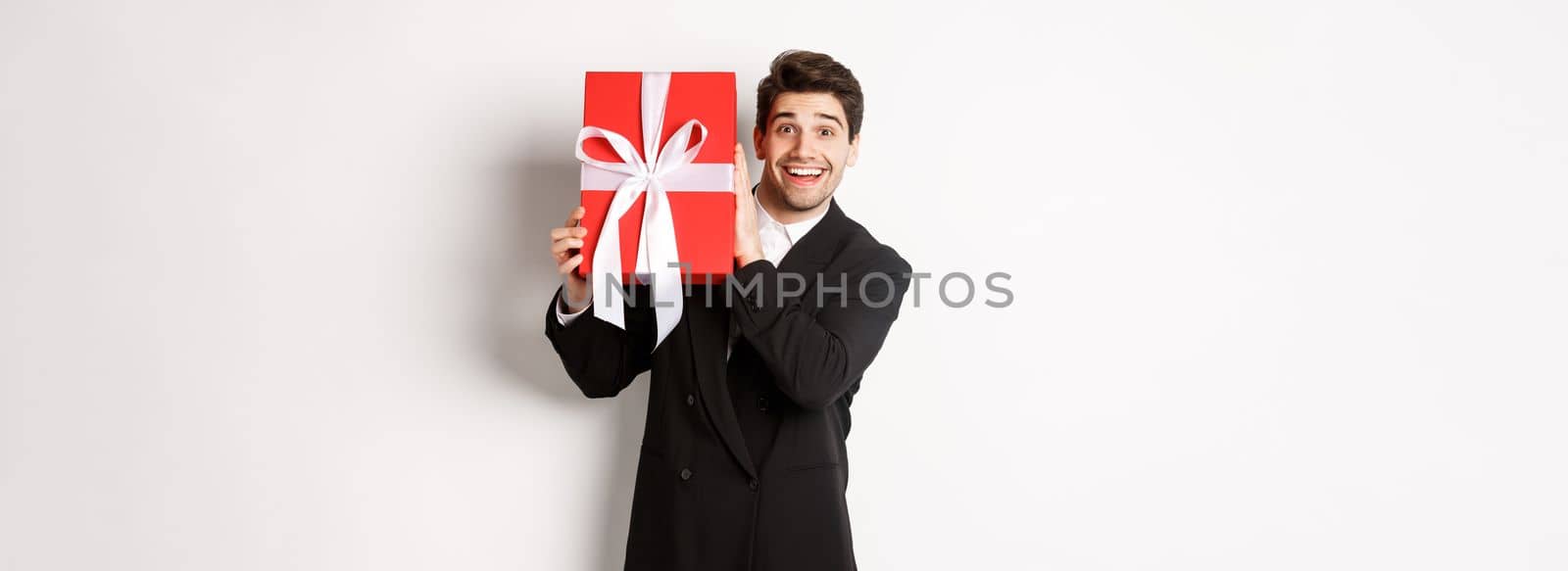 Handsome man in black suit, receiving christmas gift, smiling amazed, standing against white background.