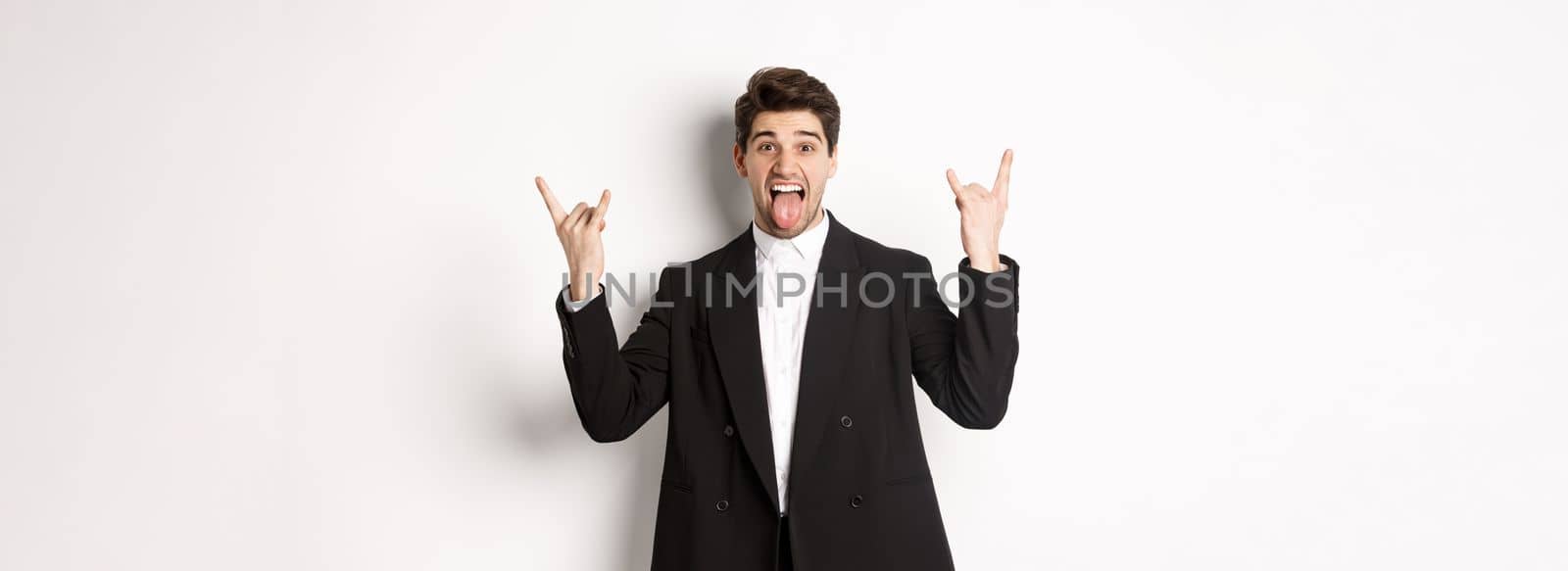 Portrait of happy attractive guy having fun at party, wearing black suit, showing rock-n-roll sign and tongue, standing excited against white background.