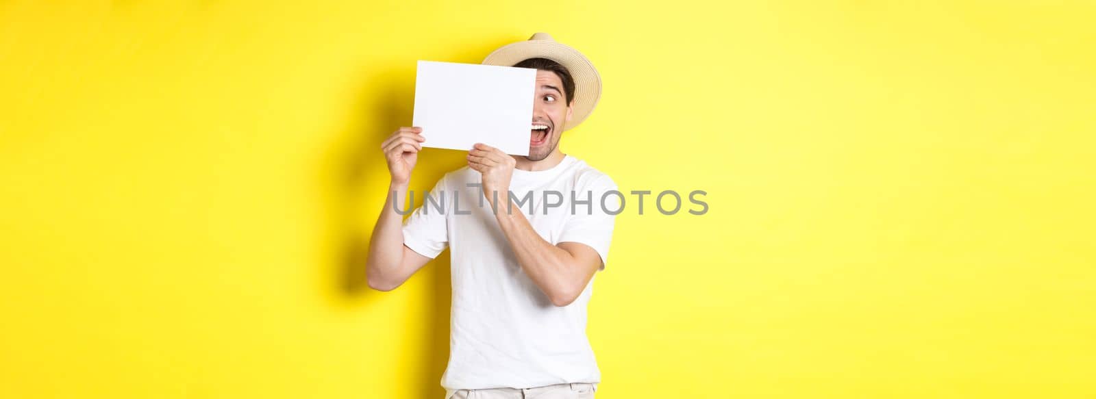 Excited man on vacation showing blank piece of paper for your logo, holding sign near face and smiling, standing against yellow background.