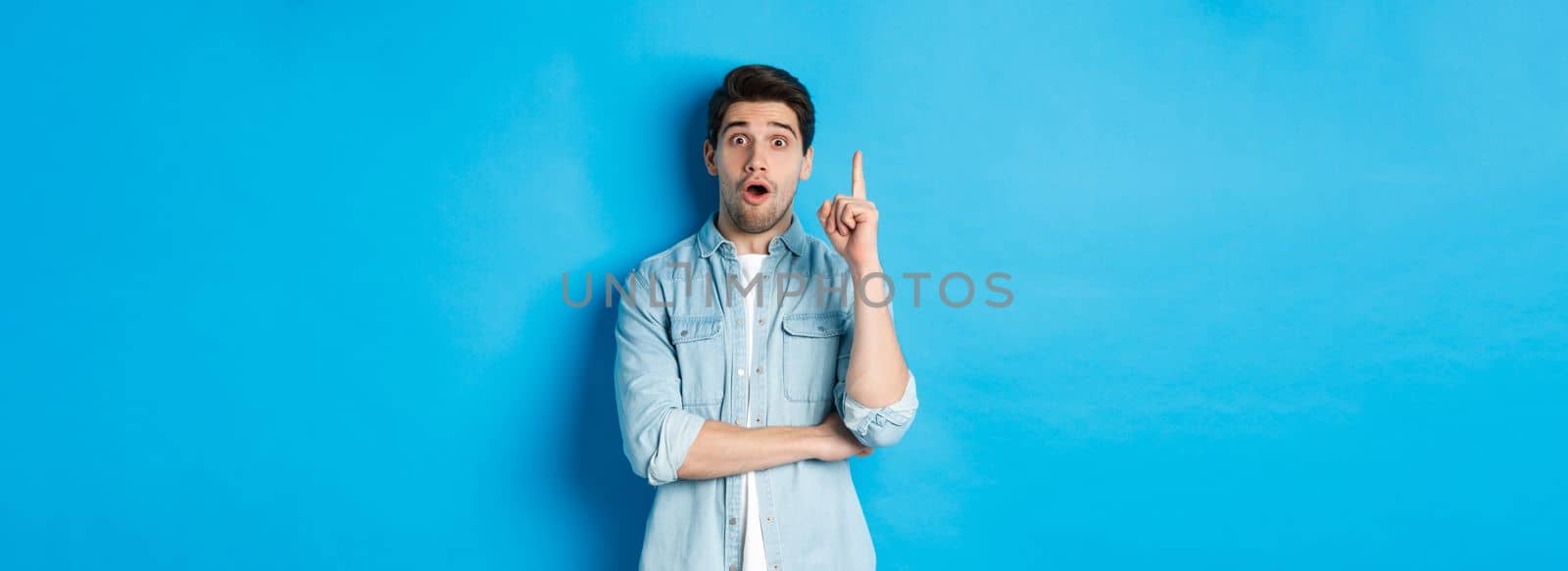 Image of handsome adult man having an idea, raising finger and smiling excited, found solution, standing against blue background.