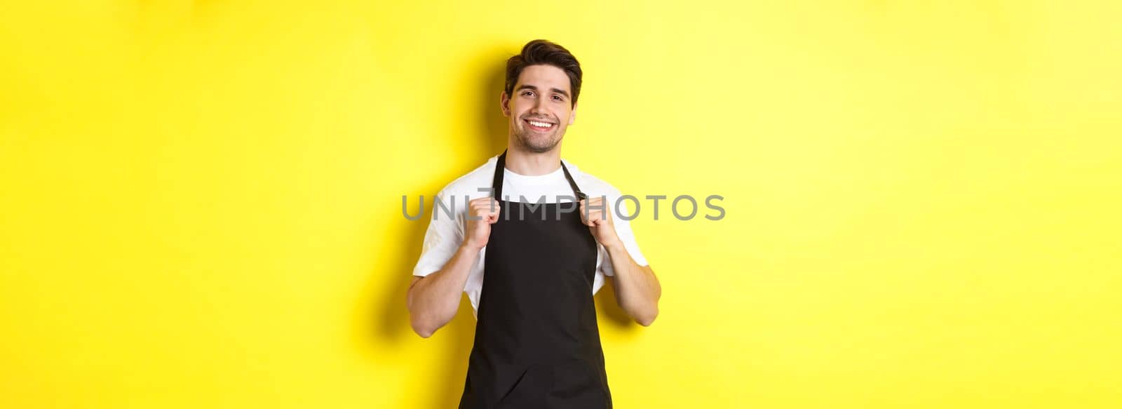 Confident barista in black apron standing against yellow background. Waiter smiling and looking happy.