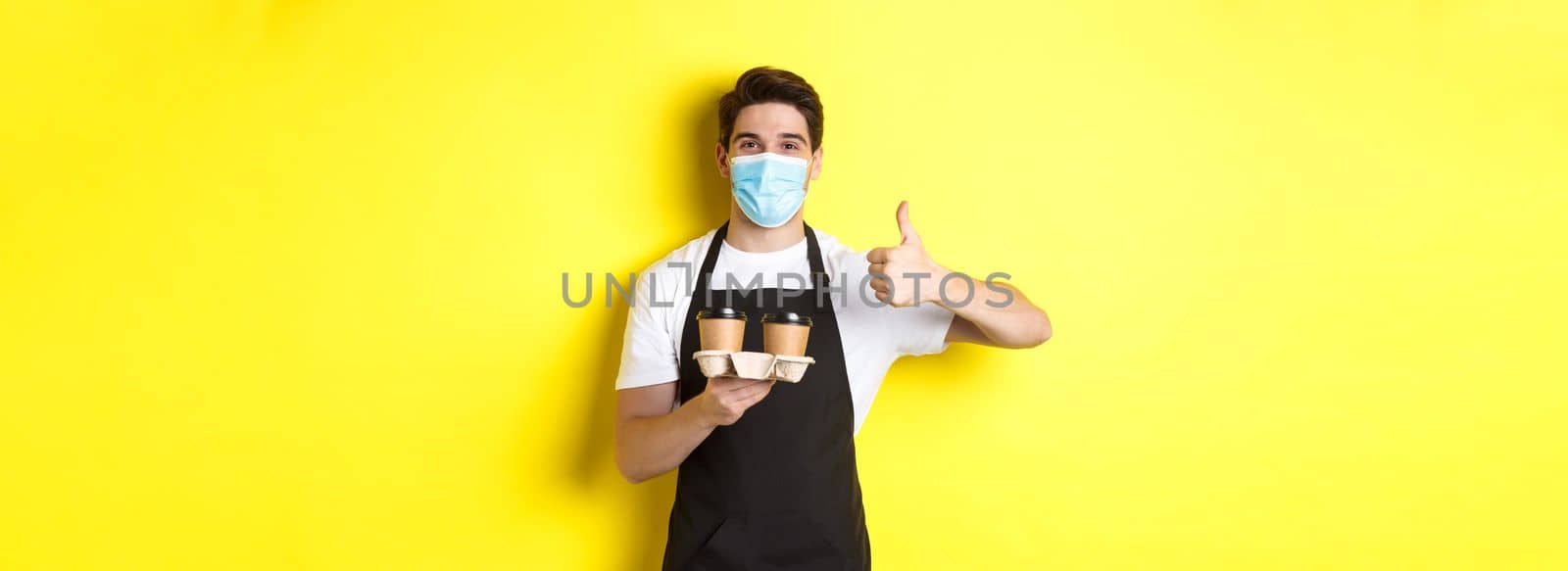 Concept of covid-19, cafe and social distancing. Young male barista in medical mask and black apron holding takeaway coffee cups, showing thumb up, yellow background.