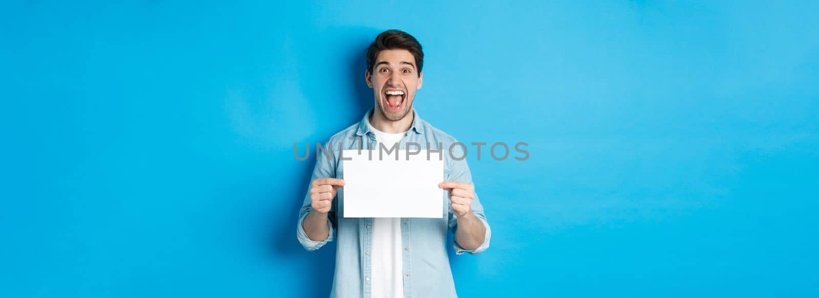 Happy attractive man showing piece of paper for your logo sign, standing amazed against blue background.