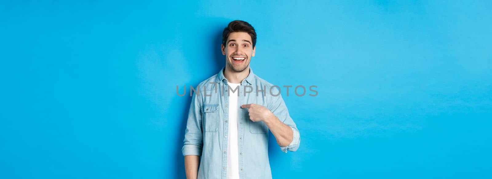 Happy and surprised man pointing at himself, smiling pleased, standing against blue background.