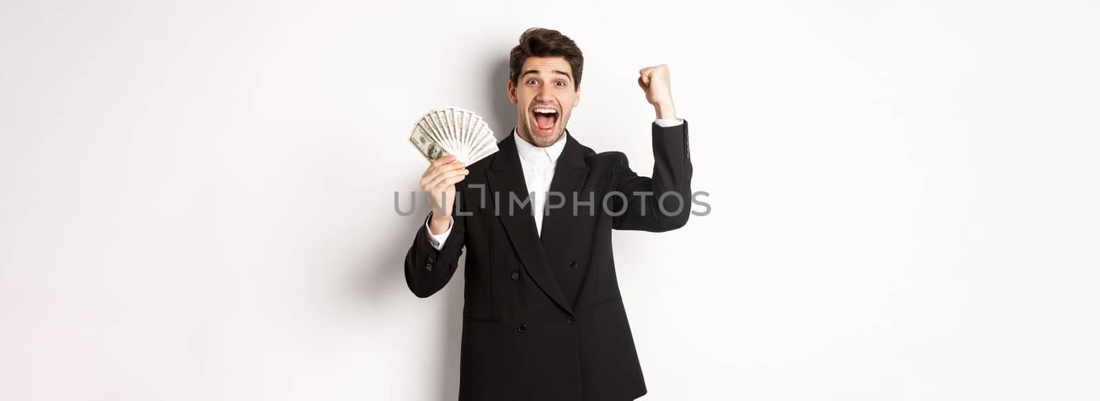 Portrait of handsome businessman in black suit, winning money and rejoicing, raising hand up with excitement, standing against white background.