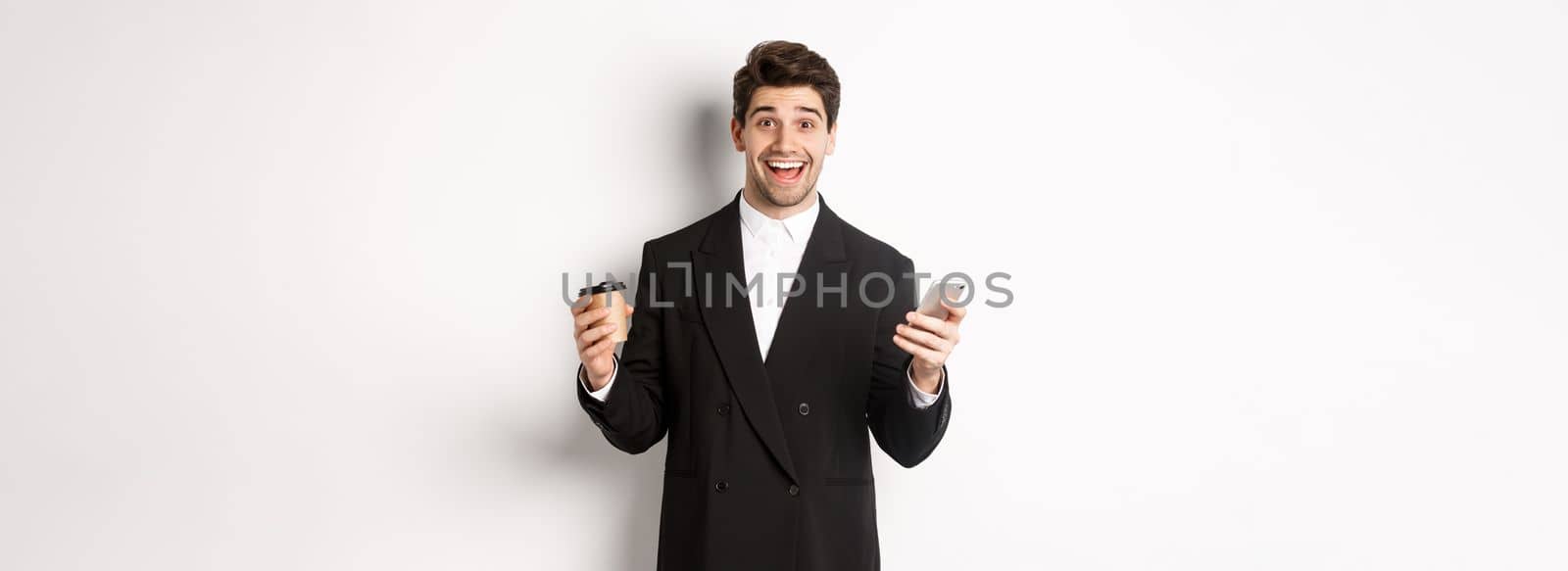 Portrait of happy good-looking man in suit, holding cup of coffee and smartphone, achieve app goal, standing over white background.