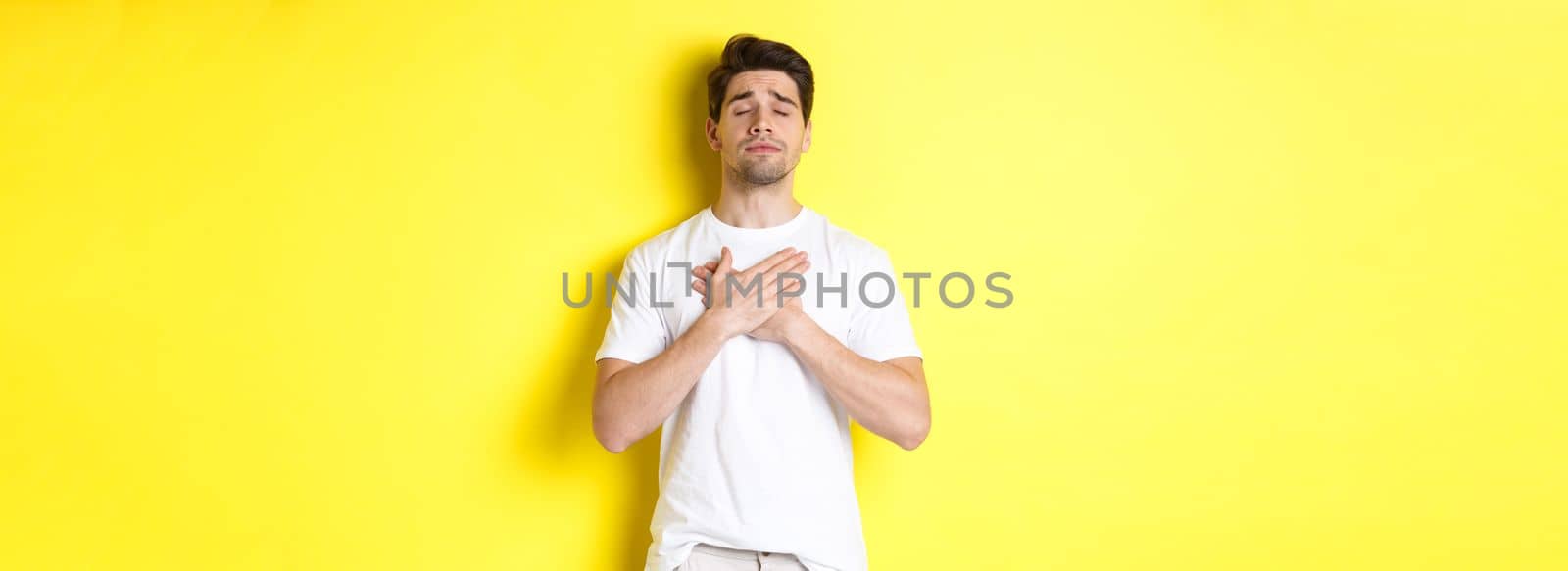 Romantic and nostalgic man holding hands on heart, close eyes and remember something, standing over yellow background.