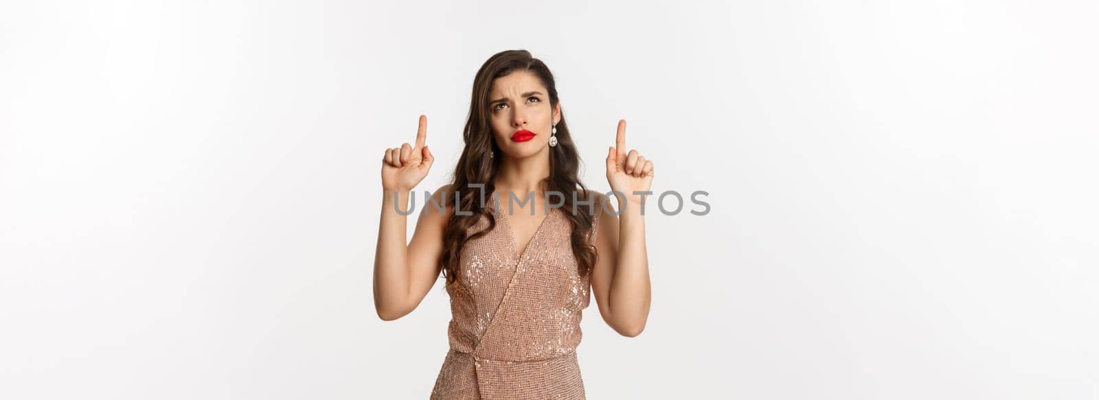 Christmas, holidays and celebration concept. Elegant woman in luxury dress looking doubtful at logo, pointing fingers up, standing over white background.