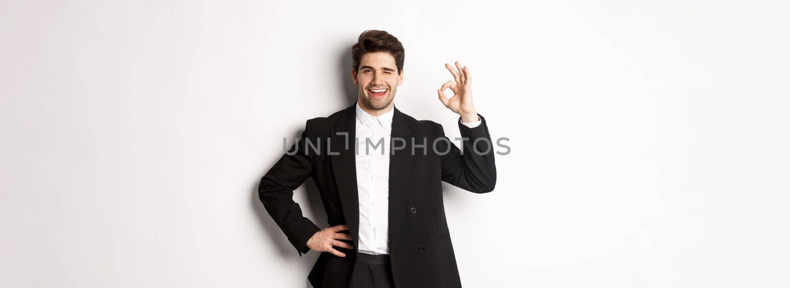 Concept of new year party, celebration and lifestyle. Portrait of confident, successful businessman in suit, showing okay sign and winking, approve something good, white background.