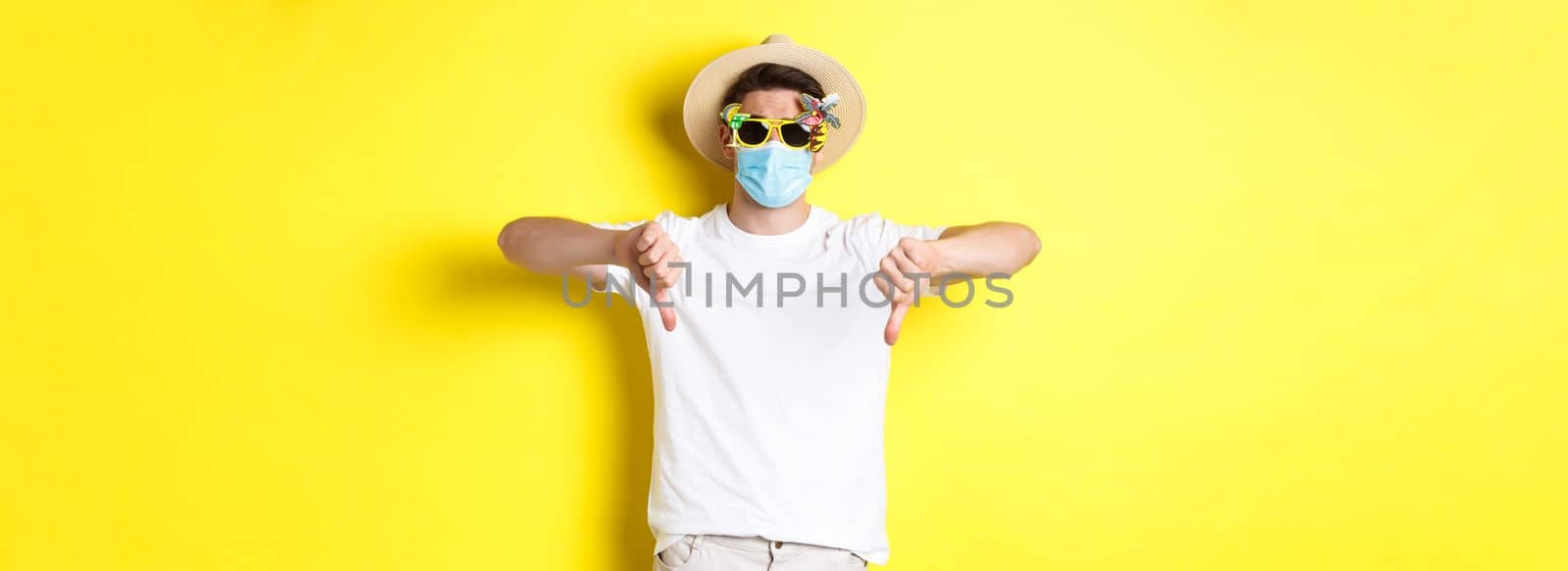 Concept of covid, vacation and tourism. Disappointed tourist complaining on lockdown during pandemic, wearing medical mask and sunglasses, showing thumbs down.