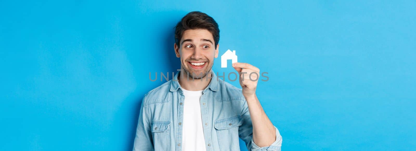 Real estate concept. Excited guy looking at small house model and smiling, renting apartment, standing over blue background.