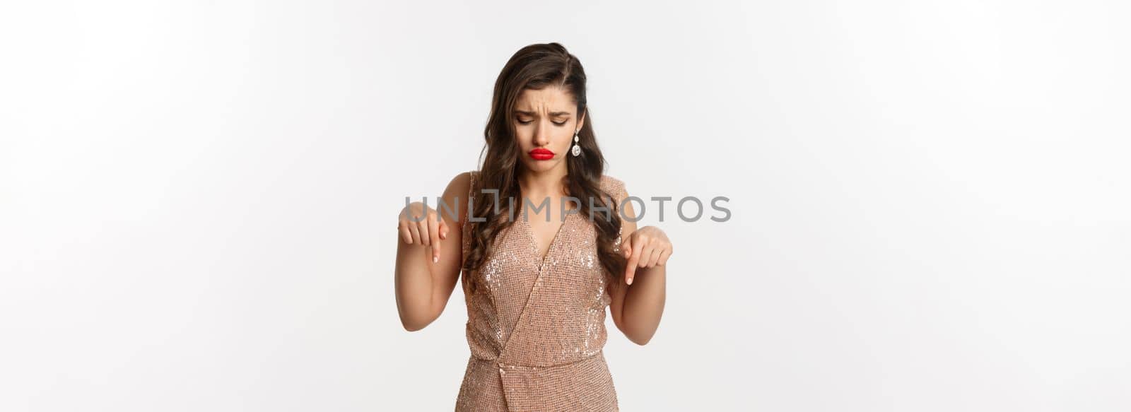 Sad beautiful girl pointing fingers down and sulking, wearing dress for christmas party, looking disappointed, standing over white background.