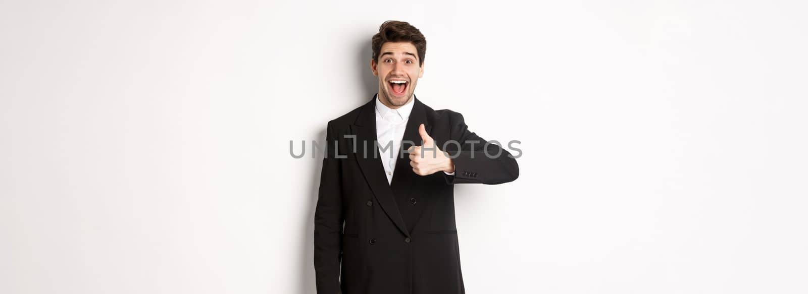 Portrait of amazed and impressed handsome man in black suit, showing thumb-up, praise awesome product, liking something good, standing against white background.