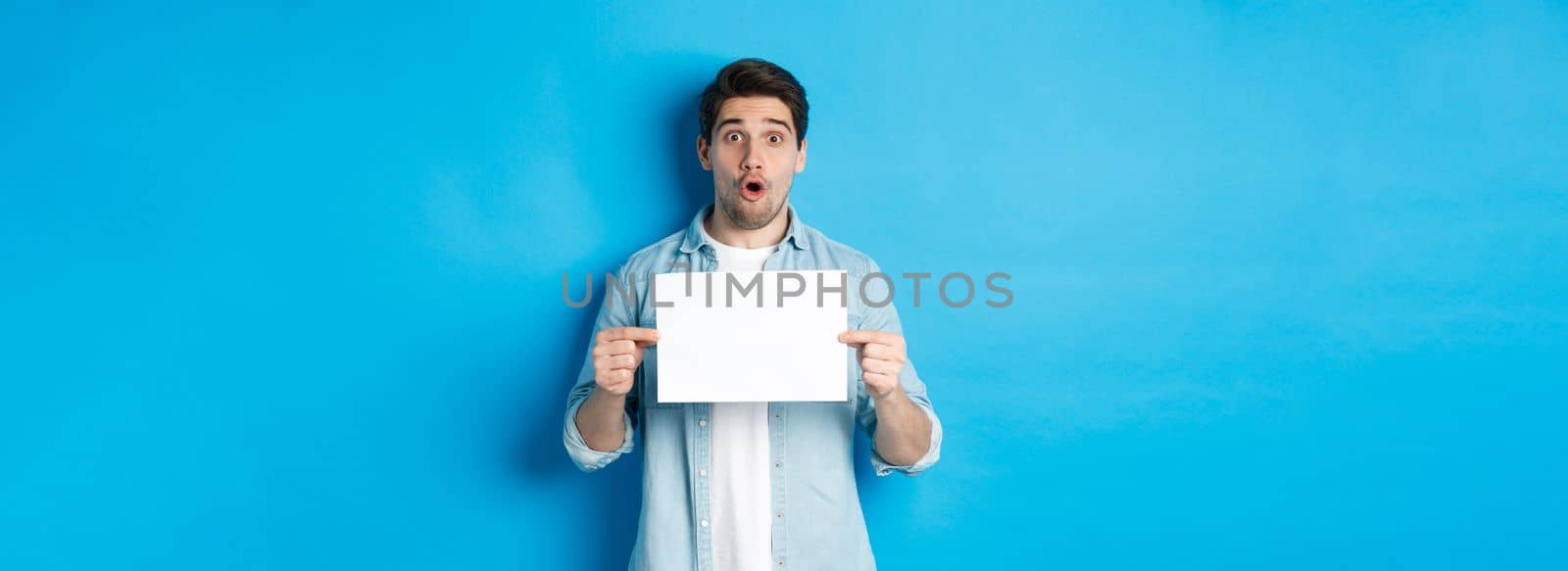Surprised man gasping and looking impressed at camera, showing blank piece of paper for your sign, standing over blue background.